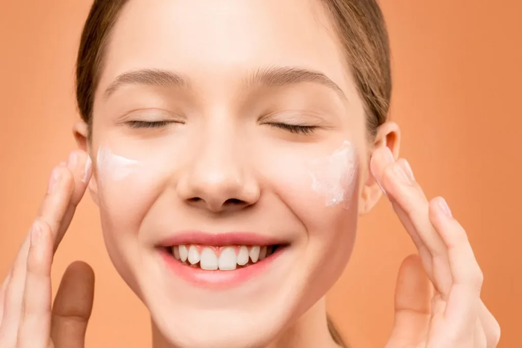 Smiling woman with cream on her cheeks, eyes closed, against an orange background