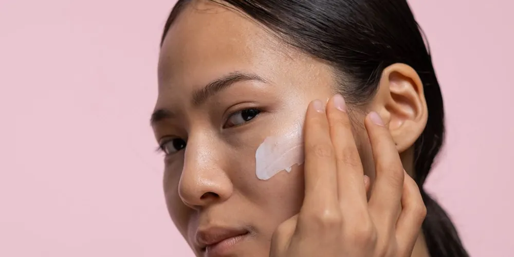Woman smoothing moisturiser onto her cheek against a soft pink background